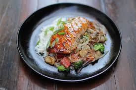 Catering 71 Ginger Soy Salmon served on a black plate with grilled salmon glazed in soy-ginger sauce, placed over stir-fried vegetables, rice noodles, and garnished with green onions.